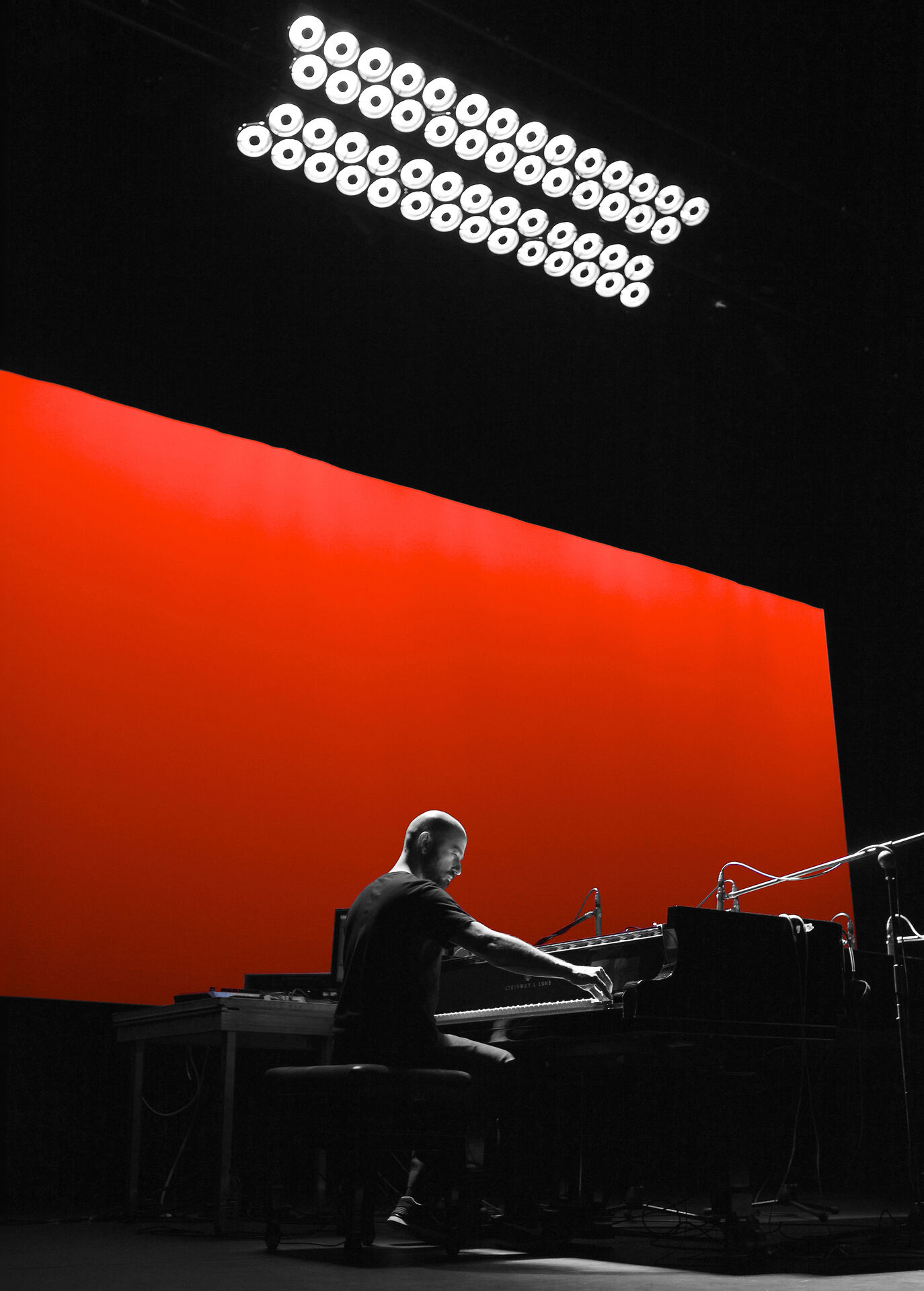 Expanded Piano II: composer Stavros Gasparatos seated at a grand piano on a dark stage, illuminated by a large red backlit panel and a grid of round white stage lights overhead.